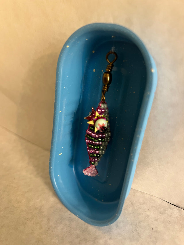 Nærbillede af håndlavet perlefisk i lilla og grønne glasperler med gul stjernekrone og pink hale, liggende i åben blå konservesdåse fra Harritsø