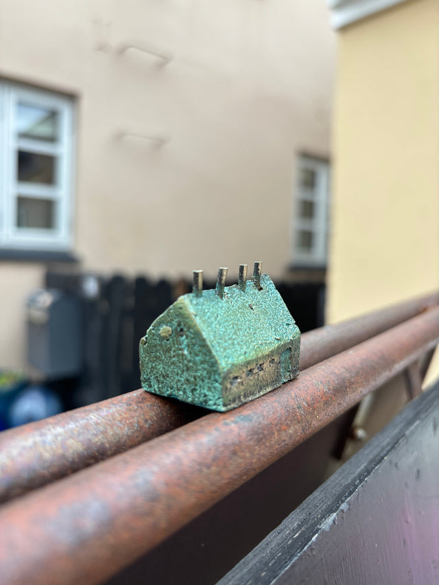 Hus #17 fra Harritsø på en rusten gelænder - bred og lav blågrøn bronze skulptur med sadeltag