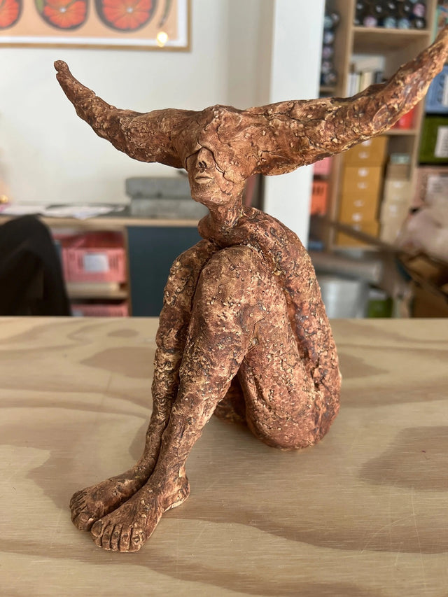 Siddende keramikskulptur med asymmetriske horn i terrakotta, set fra siden i butiksmiljø