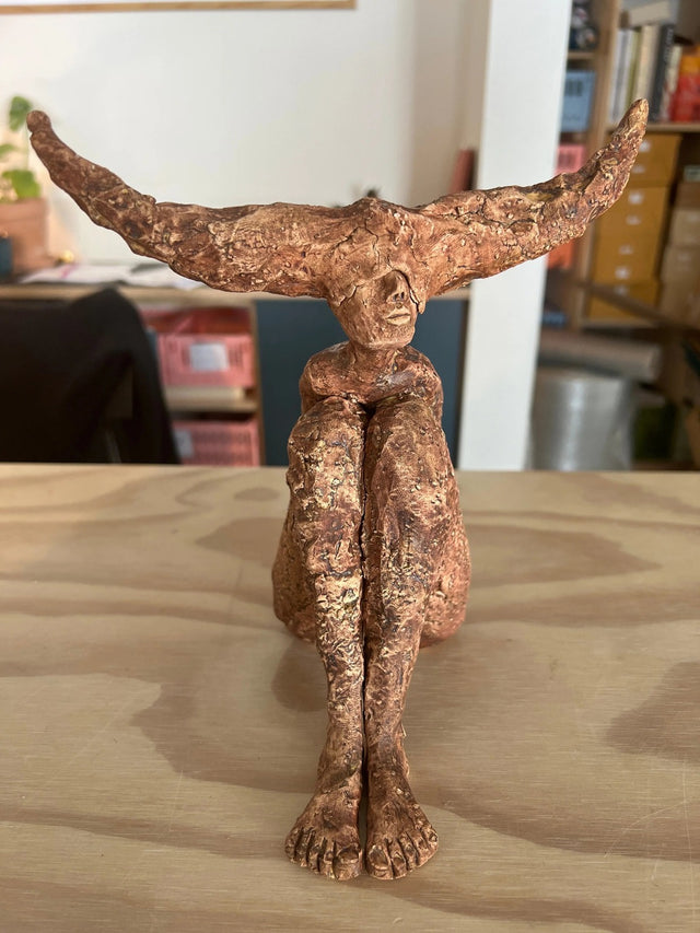 Keramikskulptur med brede horn i terrakotta set forfra, knæene trukket op mod kroppen